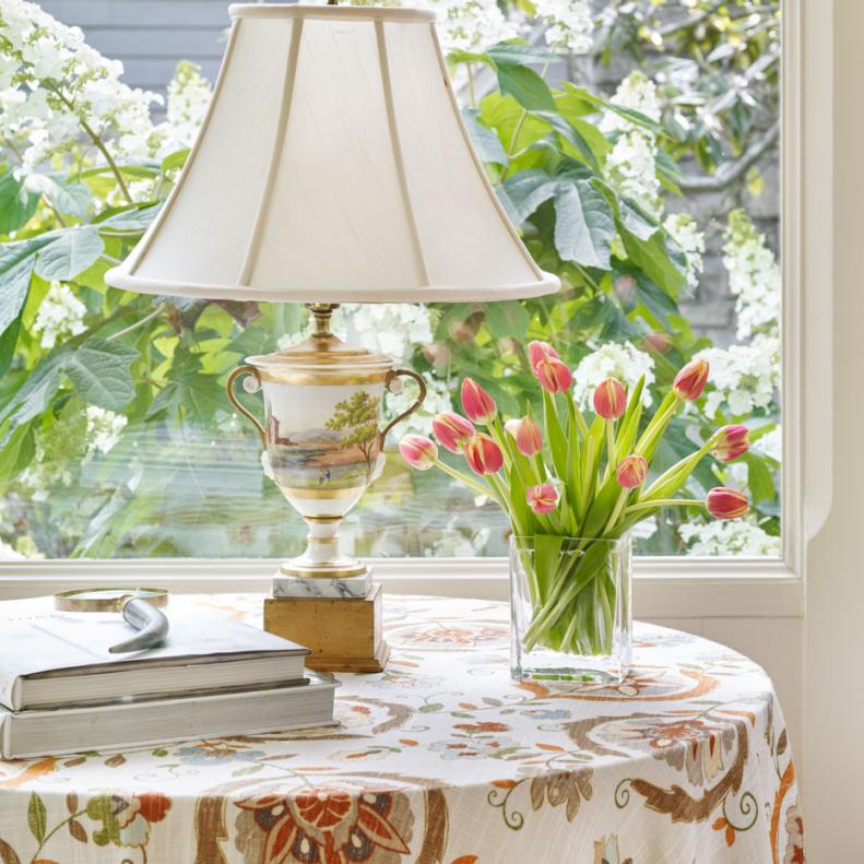 A table draped in a pretty botanical print and a glass vase of tulips perfectly frame a picture window offering a garden view just beyond.