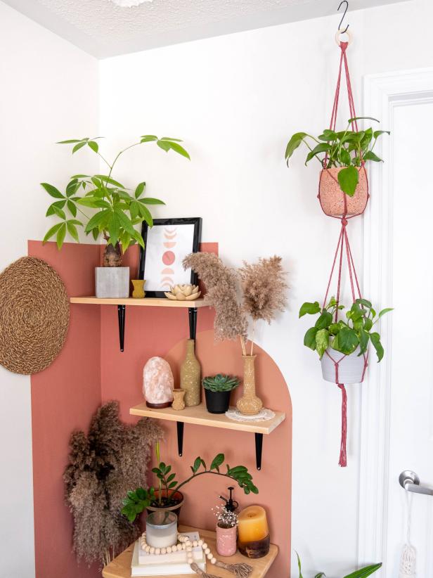 A color block painted wall on a cozy room corner with boho decor on wooden shelves. Double macrame plant hanger is hanging from the ceiling. Bohemian home accents are displayed on natural wood shelf.
