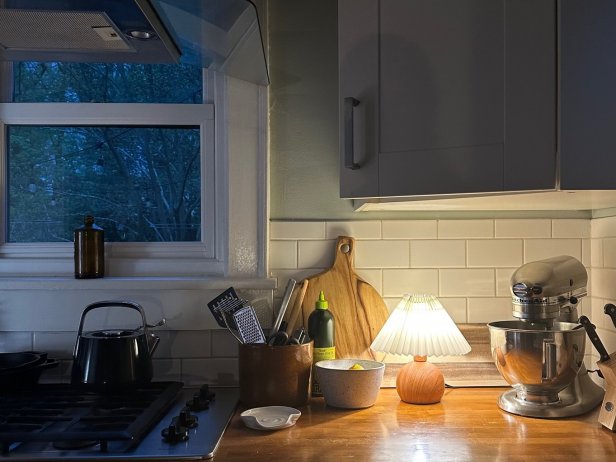 The addition of a mini table lamp, like this one with a round wood base and cone-shaped pleated shade, to a kitchen counter can add a soft, cozy glow to the space.