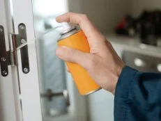 closeup of a caucasian man, in blue overalls, spraying some lubricating oil on the hinge of a white door, in a panoramic format to use as web banner or header