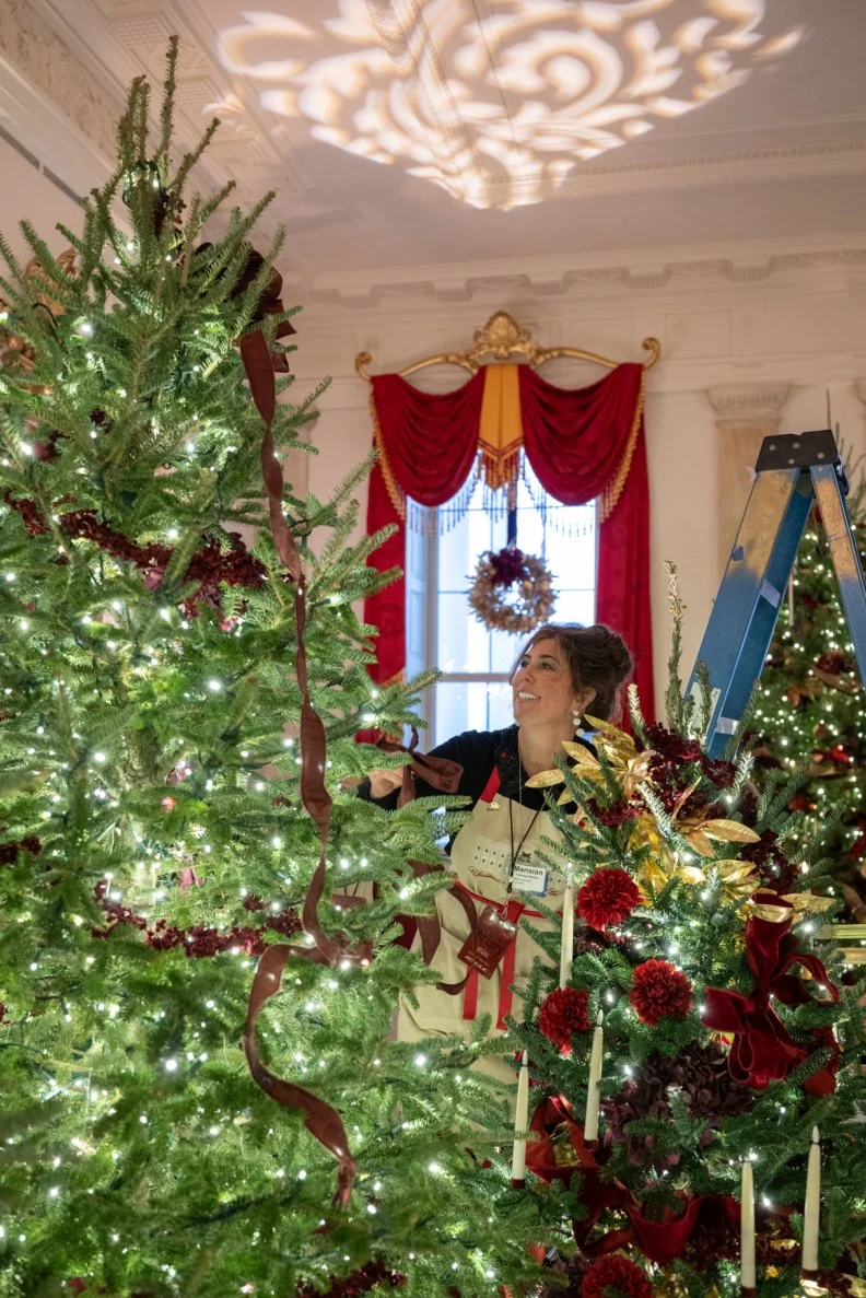 A volunteer is flanked by charming red flowers, curtains, and dramatic lengths of burgundy ribbon in the Grand Foyer.