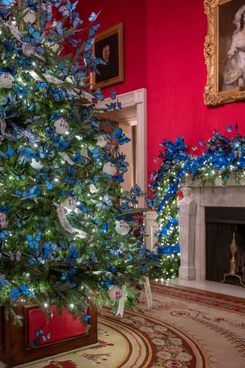 The spectacular shades of white and royal blue contrast beautifully with the brilliant Pompeiian red walls in red twill satin of this famous room. The mantle is draped with garlands interwoven with the blue butterflies that pay tribute to America's foster care community The drapery in the room was woven in the U.S. from French Empire designs. The room includes a portrait of First Lady Dolley Madison.