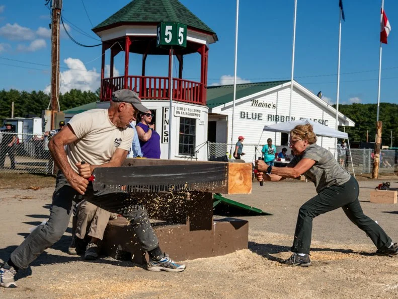 A man and a woman sawing a block of wood with a bow saw on Woodmen's Field Day at Maine's Fryeburg Fair.