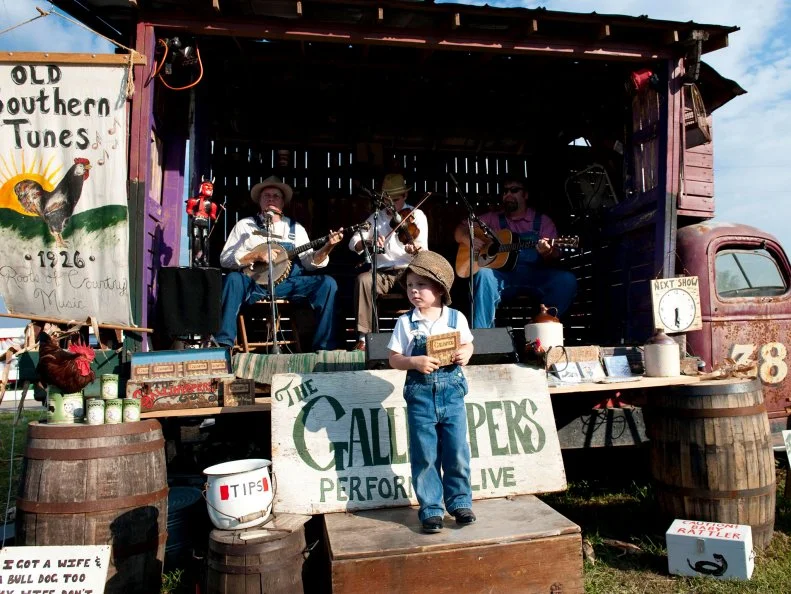 A boy in overalls and a floppy hat performing onstage with people playing banjos and violins in the background.