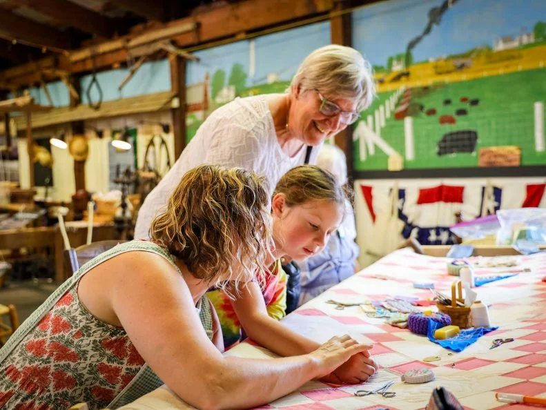An older woman bent over a younger woman and a youth, showing them how to quilt