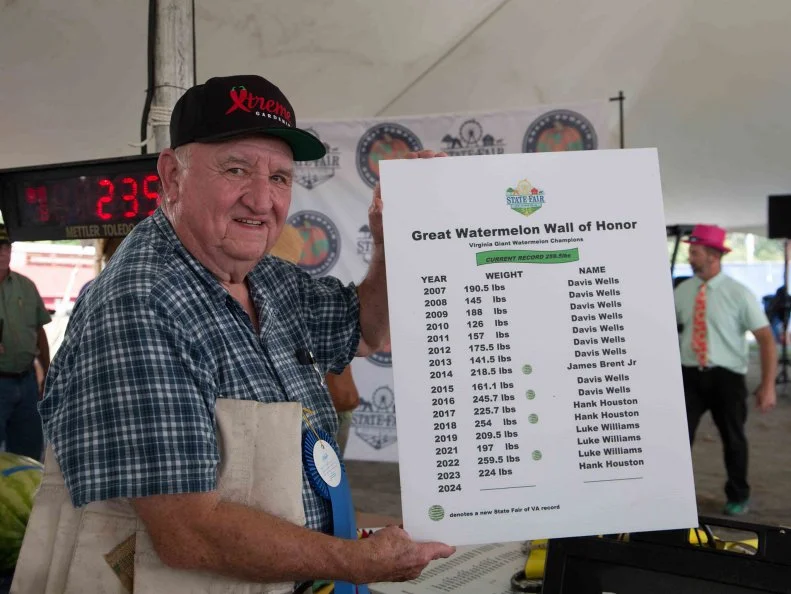 A senior man wearing a cap holding a sign listing watermelons and their weights on the Great Watermelon Wall of Honor.