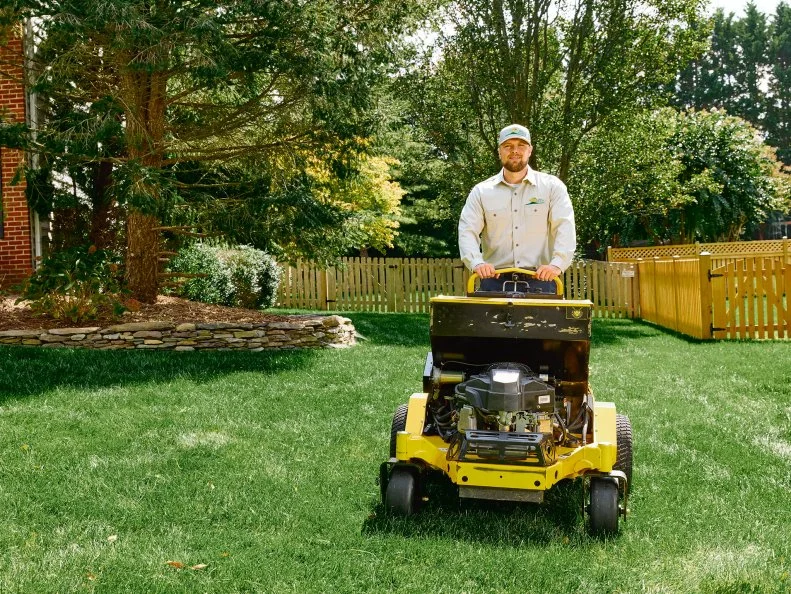 A man mowing a lawn with a fence and trees behind him