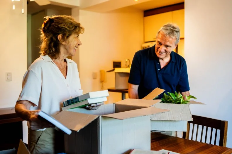 Senior couple stands by a table with open cardboard boxes, organizing belongings in a cozy home environment
