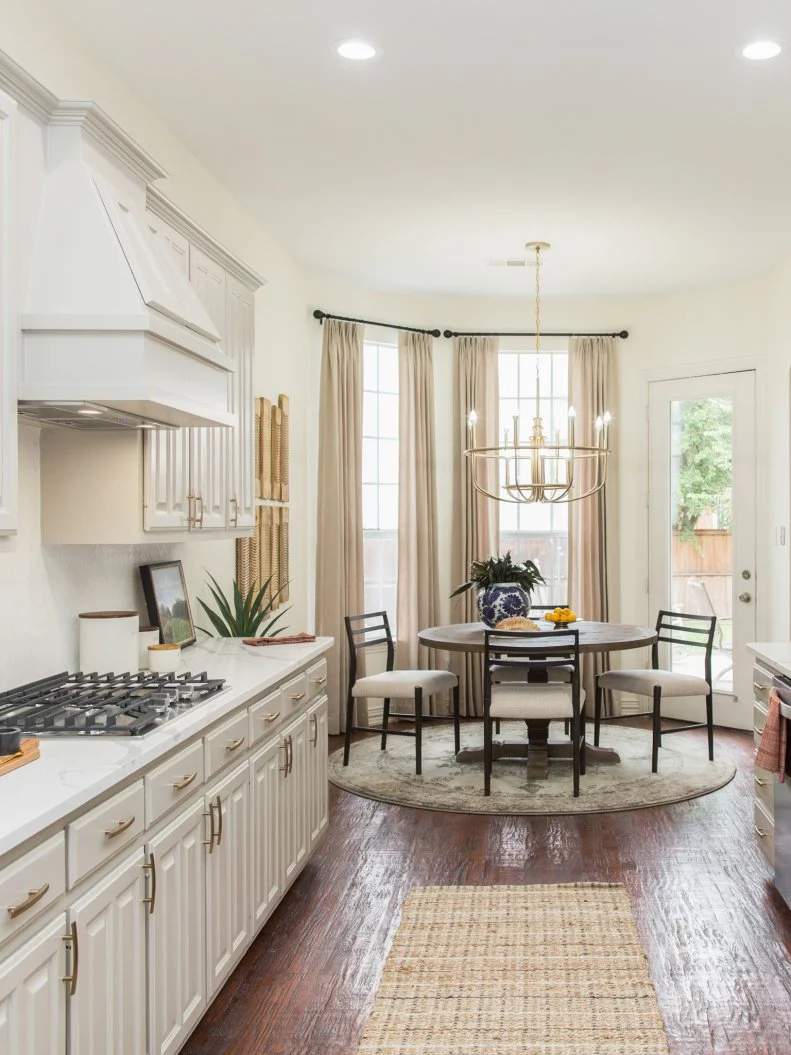 The updated kitchen in the newly renovated McNamara home has white countertops, white herringbone backsplash and bronze fixtures. The kitchen opens up to the living room, and the opening is framed by a support beam and open shelving, as seen on No Demo Reno. (after)