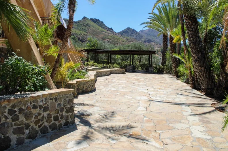 Wide shot of an empty outdoor pergola prior to makeover, highlighting wooden roof beams, tiled ground surface, iron railing, and adjacent pool in original condition, as seen on Bachelor Mansion Takeover, Season 1.