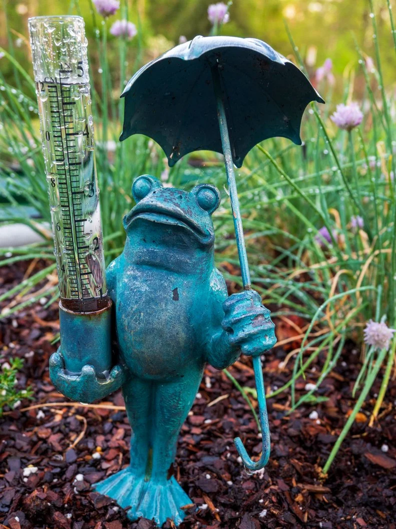 Decorative frog statue holding umbrella beside rain gauge in garden
