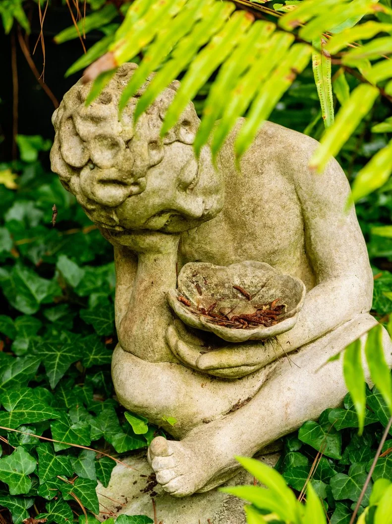 Weathered stone garden statue holding bowl beneath lush green foliage