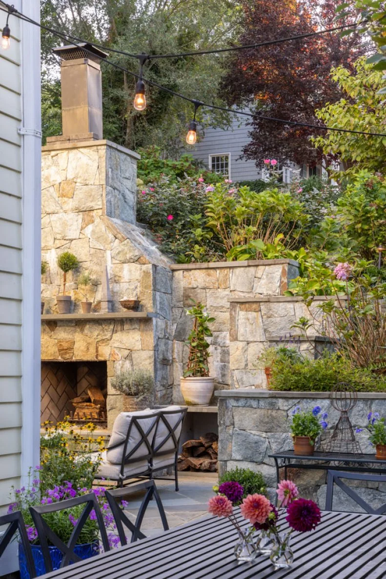 A stone patio with a fireplace, string lights overhead, terraced garden beds, and a table with vases of dahlias.