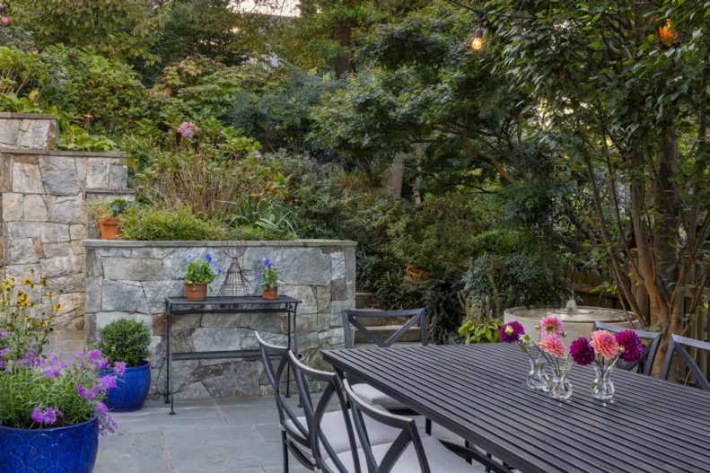 An outdoor dining table with chairs and vases of dahlias, set on a stone patio near terraced walls and a small water fountain.