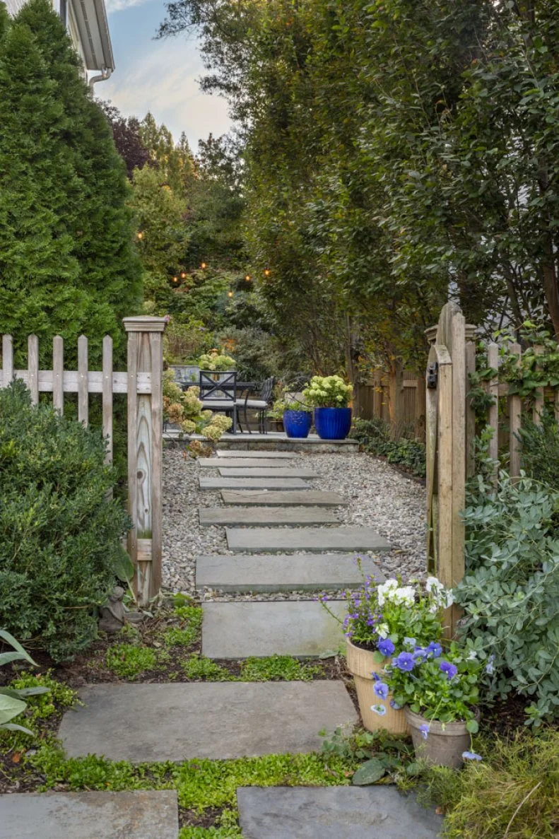A stone stepping-path through a wooden garden gate, lined with gravel and potted flowers, leading to a patio in the distance.