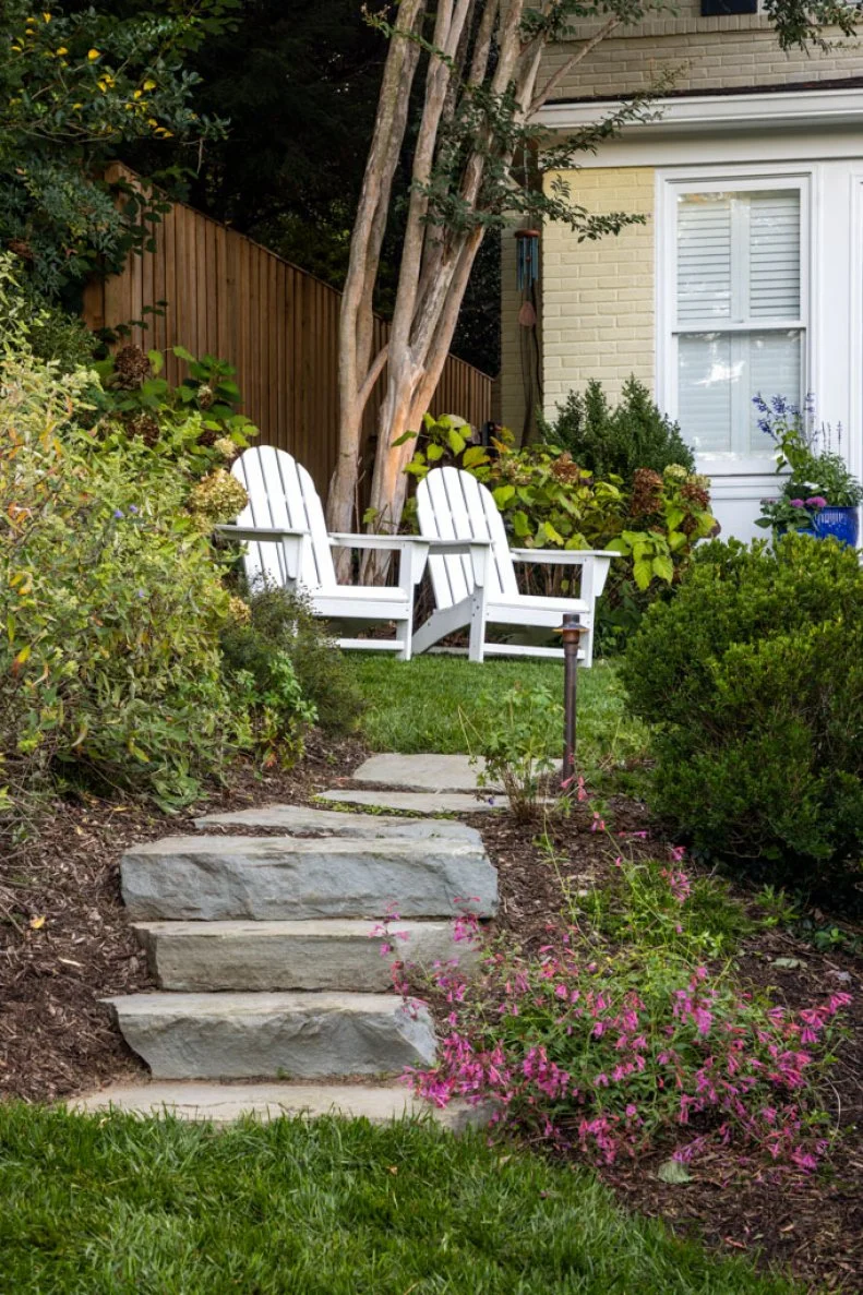 Stone steps leading up to two white Adirondack chairs surrounded by shrubs and flowering plants near a house.
