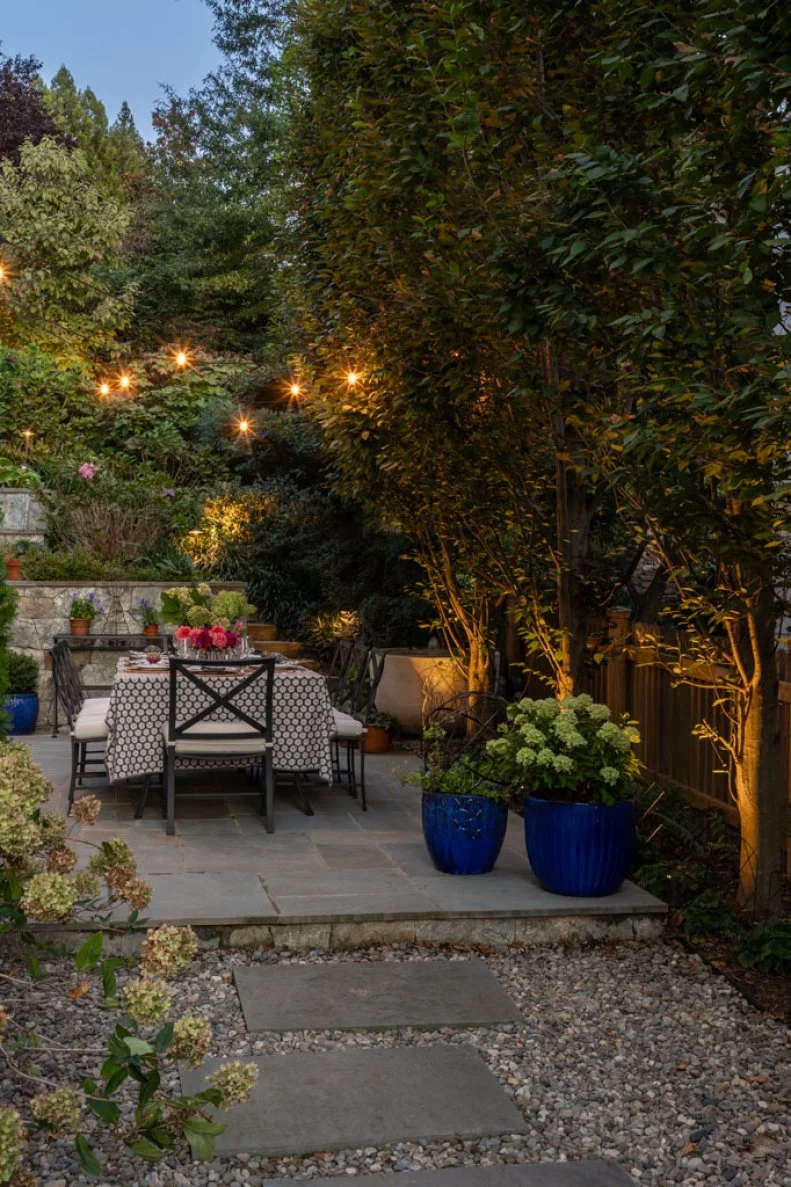 An outdoor dining table set for a meal on a stone patio at dusk, lit by string lights, with blue planters and uplit trees nearby.