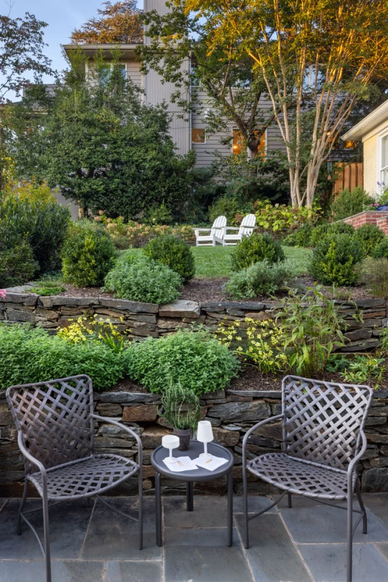 Two metal patio chairs and a small table in the foreground, with tiered stone retaining walls, boxwood shrubs, and a lawn with white chairs beyond.