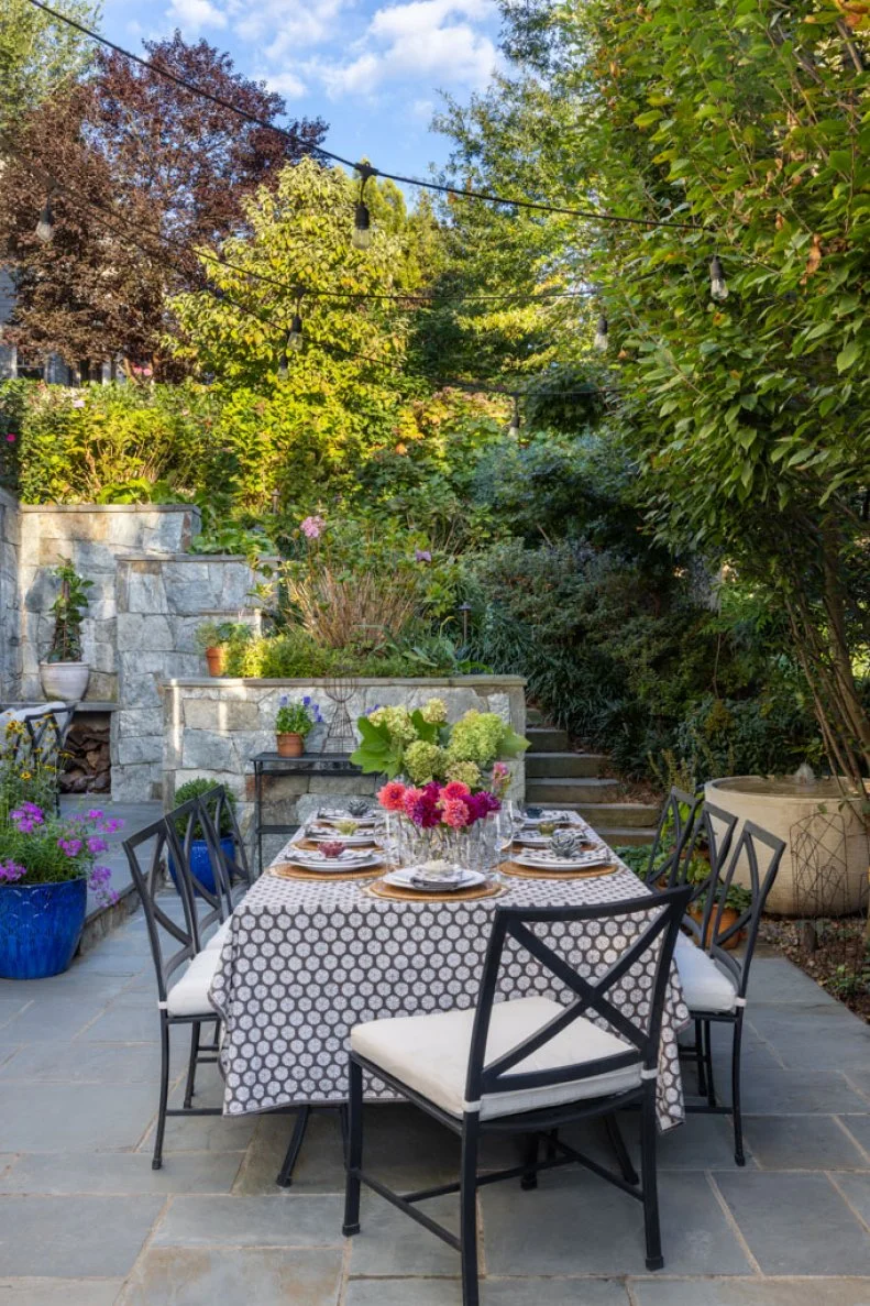 An outdoor dining table set with plates, glasses, and flowers on a patterned tablecloth, surrounded by black chairs and terraced stone walls.