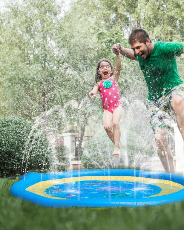 Father holding daughters hand while she jumps through the sprinkler in the garden