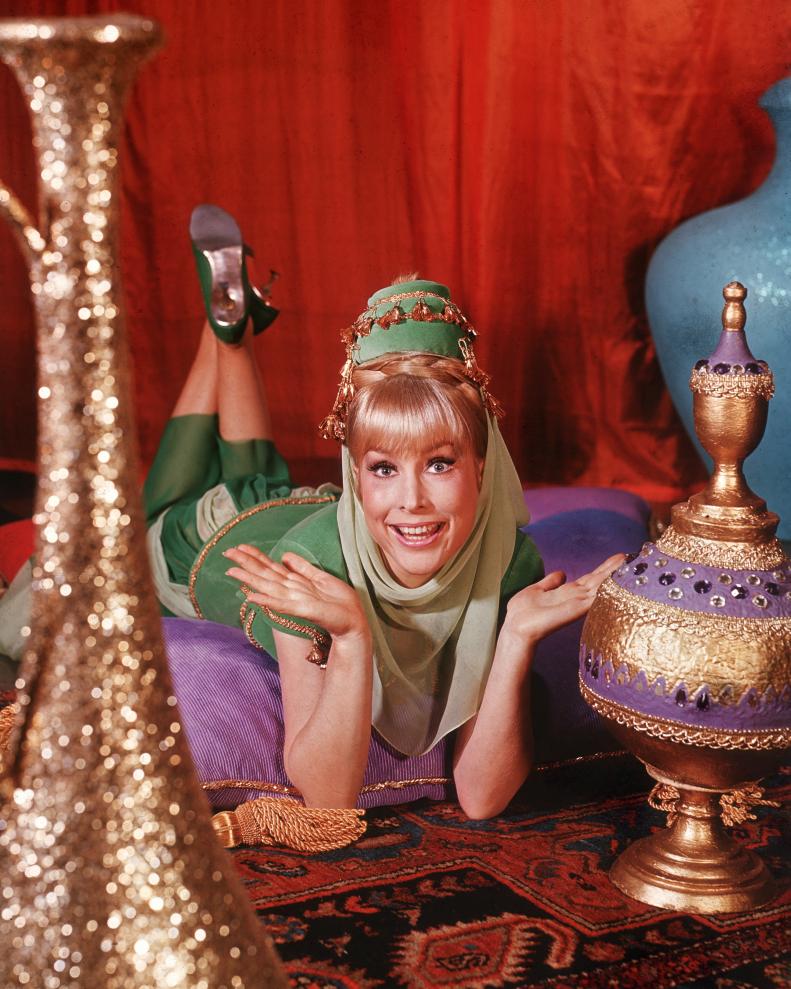 1965:  American actor Barbara Eden holds her hands up and smiles while lying on her stomach on top of cushions on an oriental rug, in a promotional portrait for the television series 'I Dream of Jeannie'. Eden is dressed in a green genie costume. Colorful vases surround her.  (Photo by Hulton Archive/Getty Images)