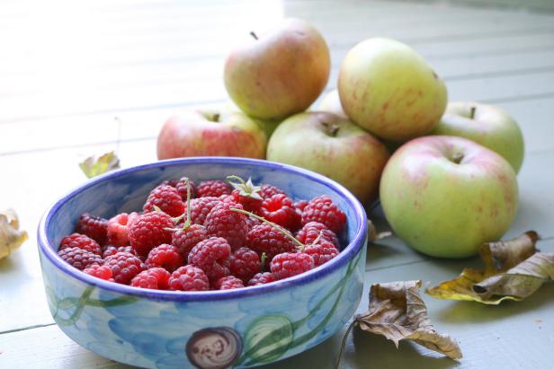Raspberries in a blue bowl with a pile of apples nearby, surrounded by autumn leaves on a green wooden floor.