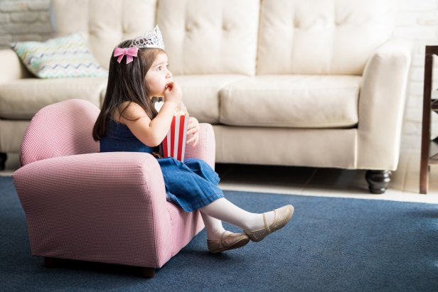 Cute baby girl wearing princess crown watching movie and eating popcorn at home