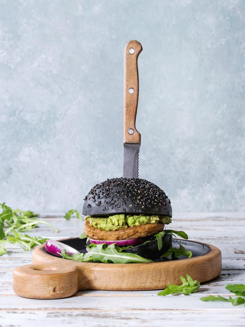 Homemade burger in black bun with avocado, arugula, onion on wood serving board over white wooden plank table. Rustic style. Homemade fast food. (Photo by: Natasha Breen/REDA&CO/UIG via Getty Images)