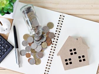 Business, finance, savings, property ladder or mortgage loan concept : Flat lay or top view of wood house model and coins scattered from glass jar on open blank notebook paper