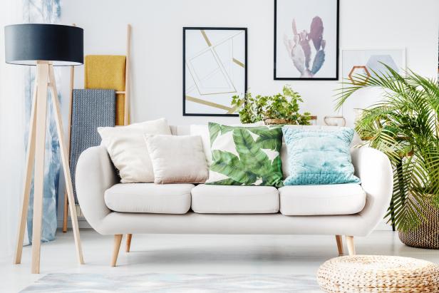 Floral cushion on a beige sofa and posters on the wall in living room with pouf, lamp and plant