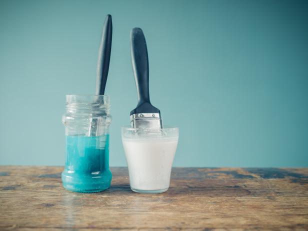 Two paintbrushes are soaking in jars of water and are coloring the water white and blue