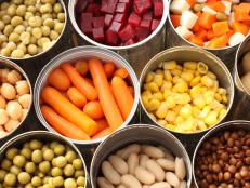 Twelve cans of open vegetables sit next to one another in three rows.  There are peas, carrots, beats, snow peas, corn, chick peas and Lima beans in the cans.