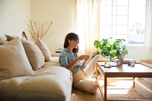 Young woman sitting on her living room floor at home wearing wireless earbuds and streaming something on a laptop