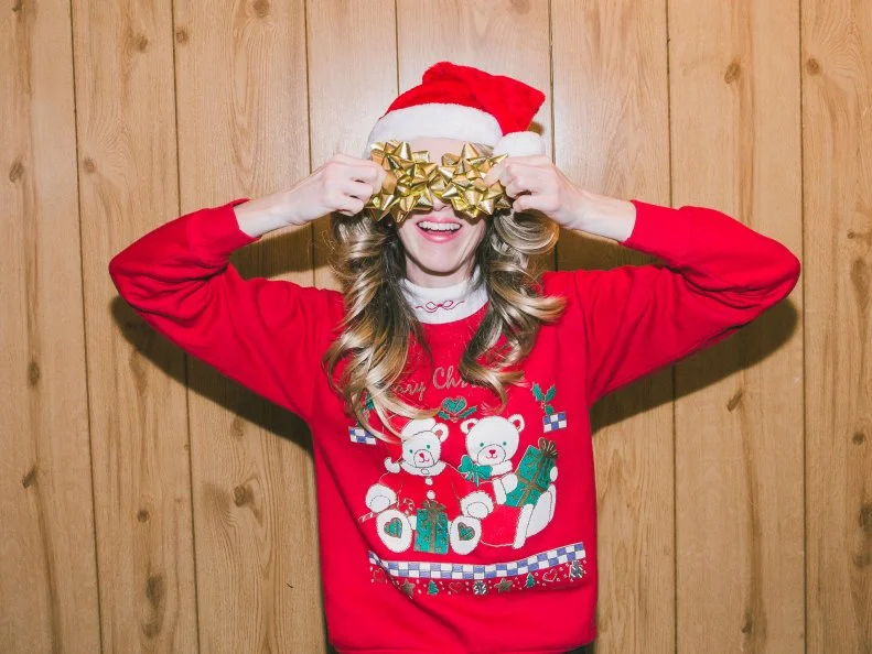 Retro woman wearing ugly Christmas sweater and Santa hat, funny Christmas woman during vintage holiday season. Happy holidays concept of cheerful woman wearing 1980s style for holiday celebration, festive winter season surprise, or secret Santa. Playful 1980s woman having fun and smiling indoors is hiding and covering eyes with gift bows inside 1980s home domestic room interior with wood paneling. Funny character with toothy smile. There is copy space on wood paneled wall. Part of a series.