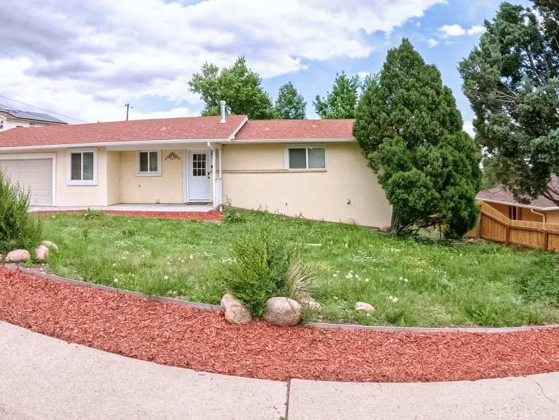 Parker, Colorado, USA-June 9, 2025 - A small beige mid-century home sits behind a yard with red mulch and minimal landscaping. Green grass and scattered rocks add contrast to the otherwise modest
