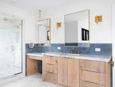 A luxurious bathroom with a wood vanity cabinet, blue tiles on the walls, gold accented lights and mirrors, and a large walk-in shower lined with marble tiles.