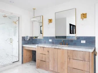 A luxurious bathroom with a wood vanity cabinet, blue tiles on the walls, gold accented lights and mirrors, and a large walk-in shower lined with marble tiles.