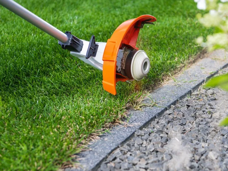 A person trims the grass close to a stone edge using a grass trimmer, ensuring a neat garden appearance.