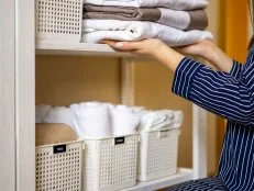 Domestic woman in pajamas neatly putting folded linens into cupboard vertical storage system use Marie Kondo method. Female housewife spring seasonal cleaning space organizer with organized boxes