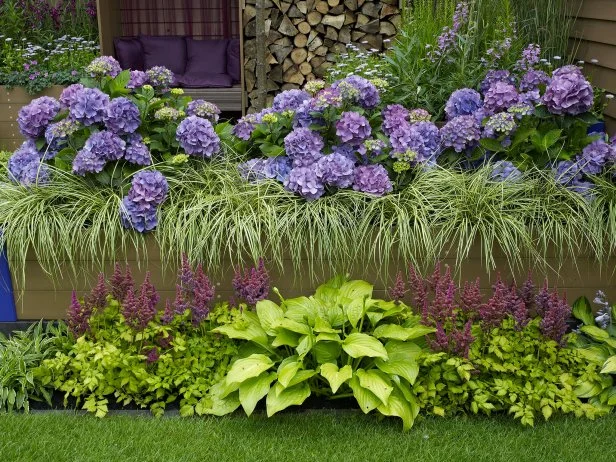 Layered raised garden bed filled with hydrangeas, ornamental grasses, and shade plants, creating a dense, colorful display in front of a cozy outdoor seating area with stacked firewood.