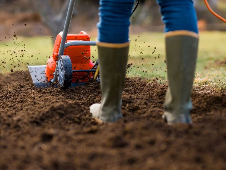 A person in rubber boots using an orange tiller to till a garden plot.
