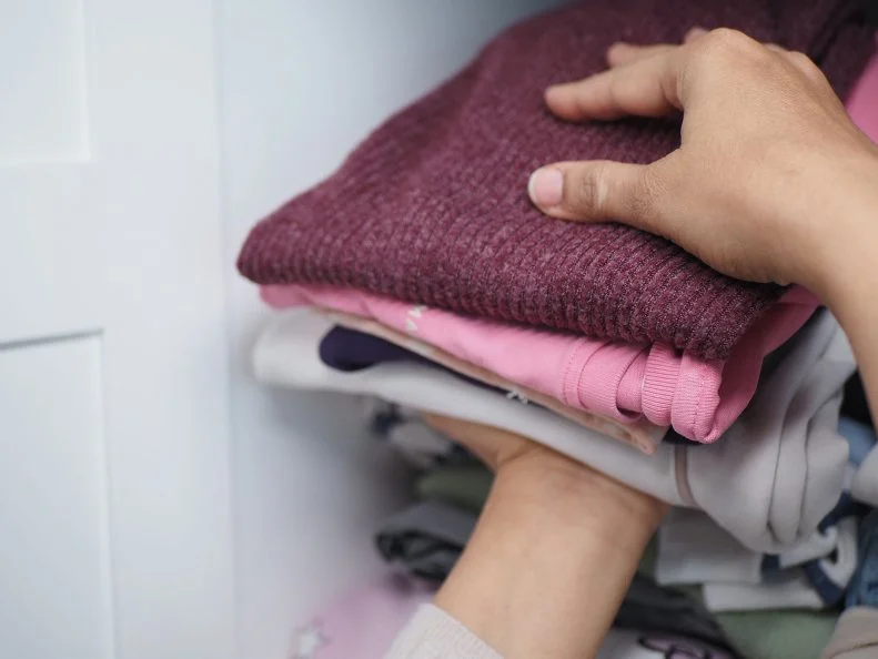 Hands placing a folded maroon sweater onto a stack of clothes on a closet shelf.