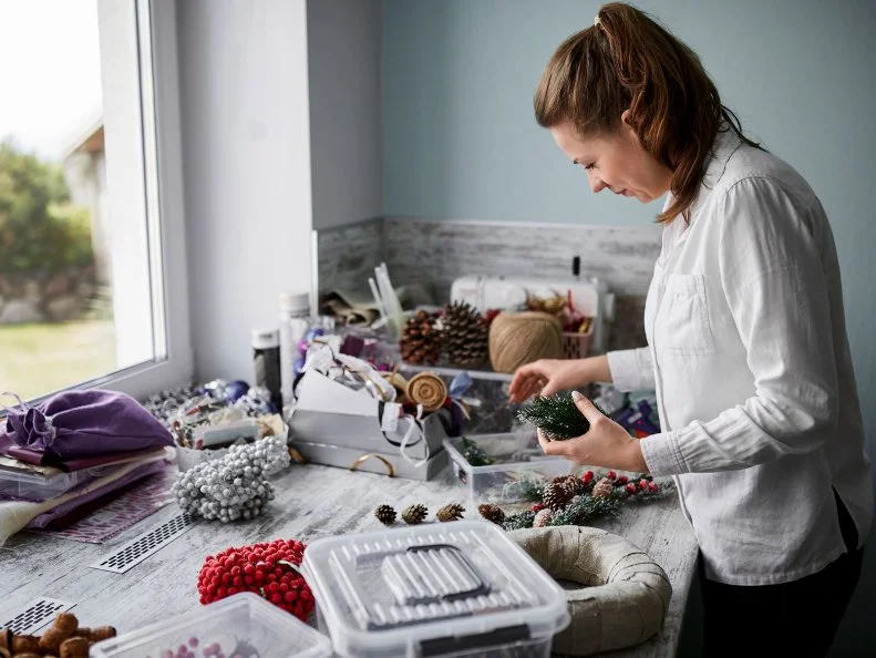 A woman sorts holiday decorations and craft supplies spread across a table.