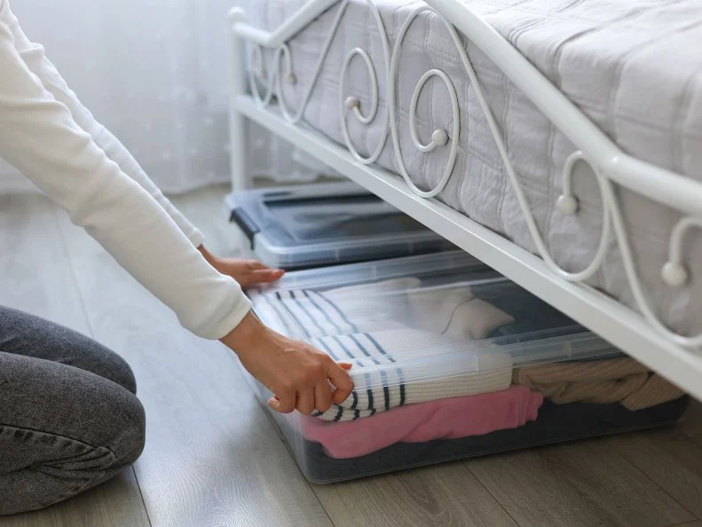 A kneeling person slides a clothing-filled storage bin out from under a bed.