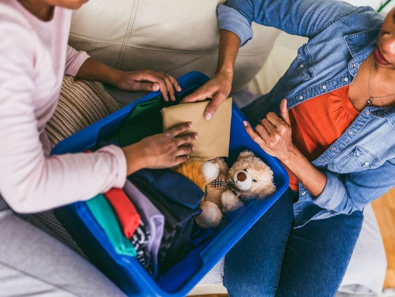 wo people sort clothes and a teddy bear into a blue bin on a couch.