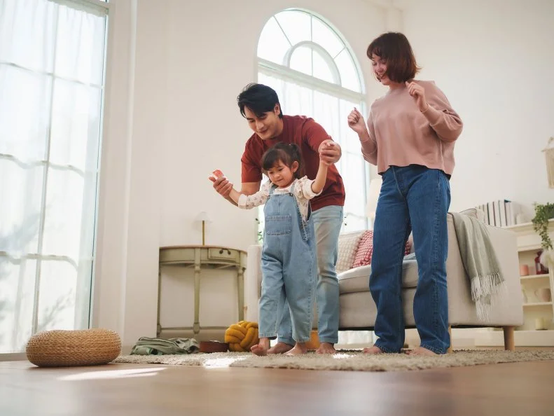 Parents dance with their young daughter in a bright, tidy living room.