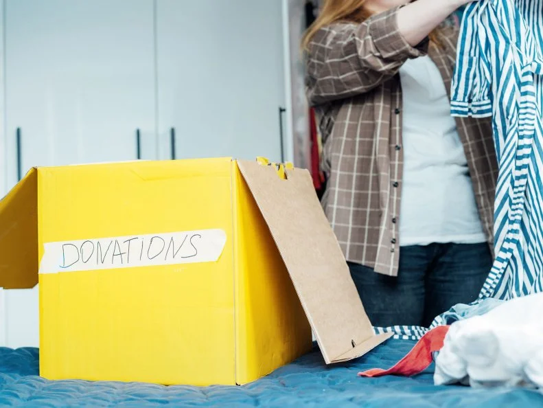A yellow box labeled "donations" sits on a bed as a woman sorts clothes in the background.