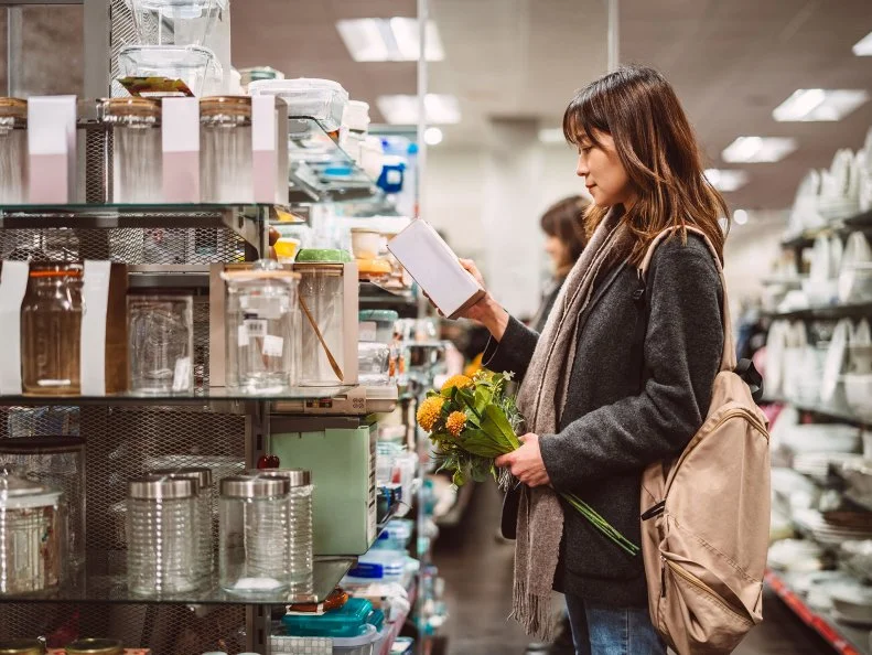A woman holding flowers reads a product box while shopping in a home goods aisle.