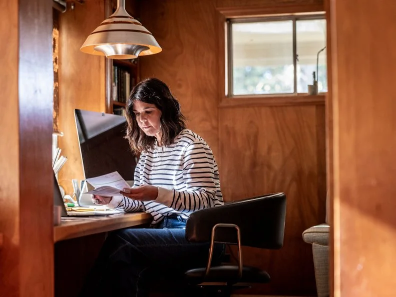 A woman reviews paperwork at a desk in a wood-paneled home office.