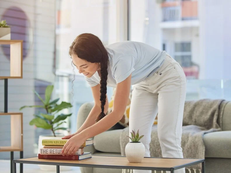 A smiling woman stacks books on a coffee table in a bright living room.